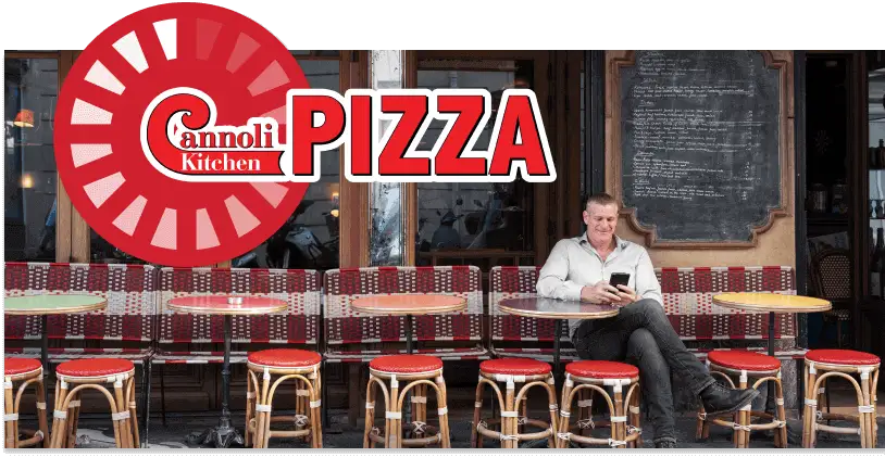 A person sits at an outdoor table of a restaurant with red chairs, the vibrant "Cannoli Kitchen" sign prominently above, hinting at delicious pizza and more.