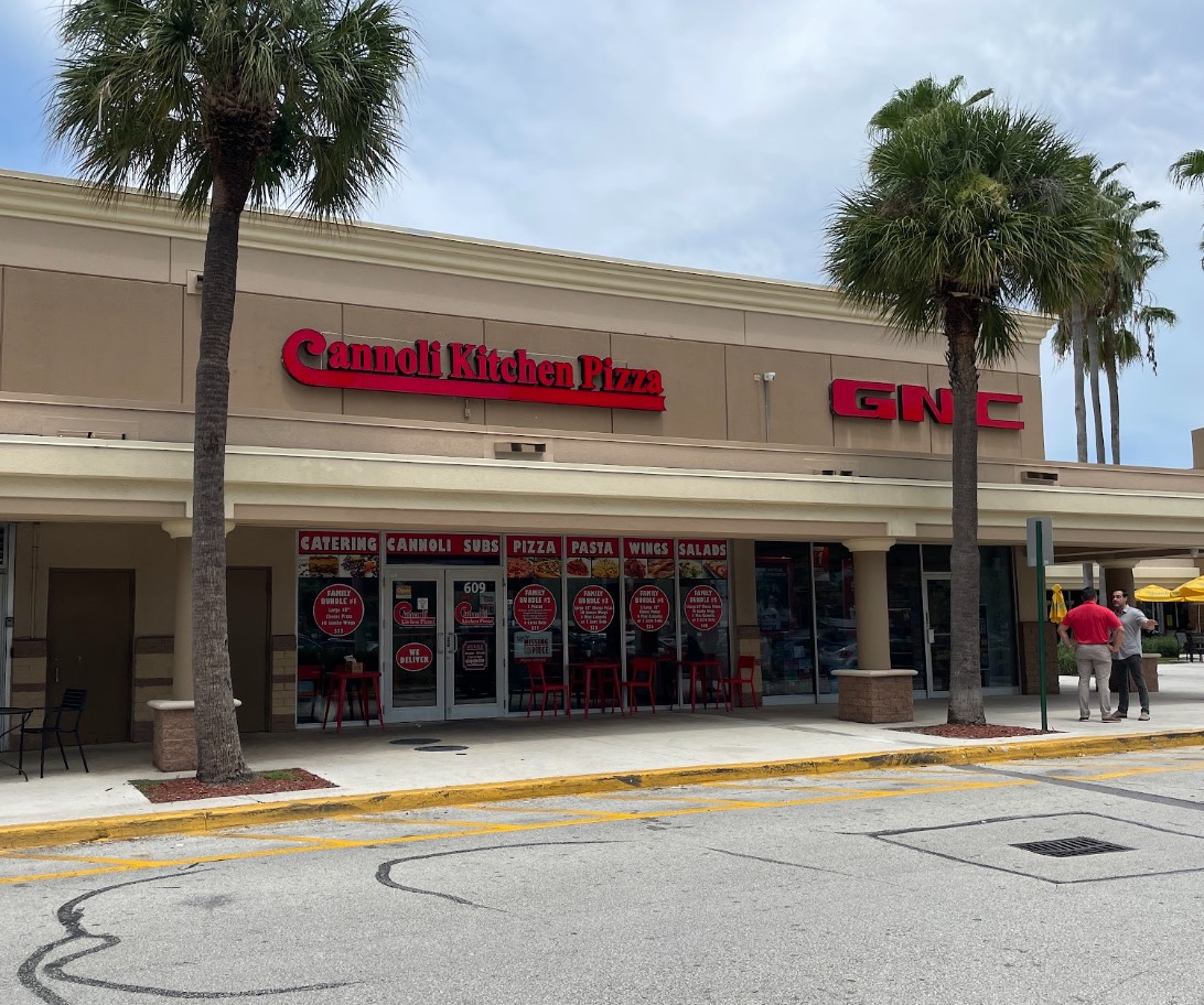 A storefront features "Cannoli Kitchen Pizza" and a GNC store in a strip mall, with palm trees and a sidewalk in front.