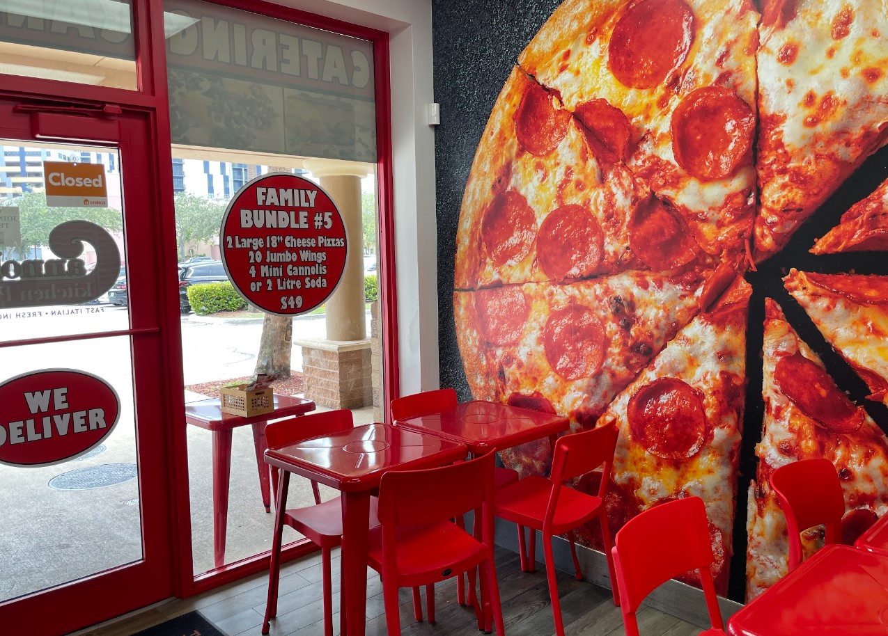 Inside a pizzeria with red chairs and tables, a large pepperoni pizza wall mural, and a window displaying delivery and family bundle options.